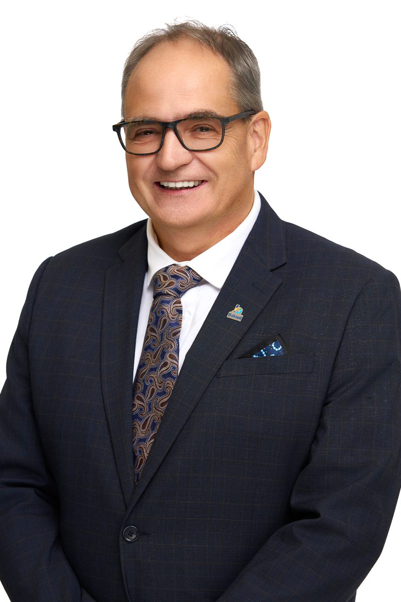 Portrait d’un homme souriant portant des lunettes foncées, un veston bleu marine à motif discret, une chemise blanche et une cravate à motifs paisley, sur fond blanc.