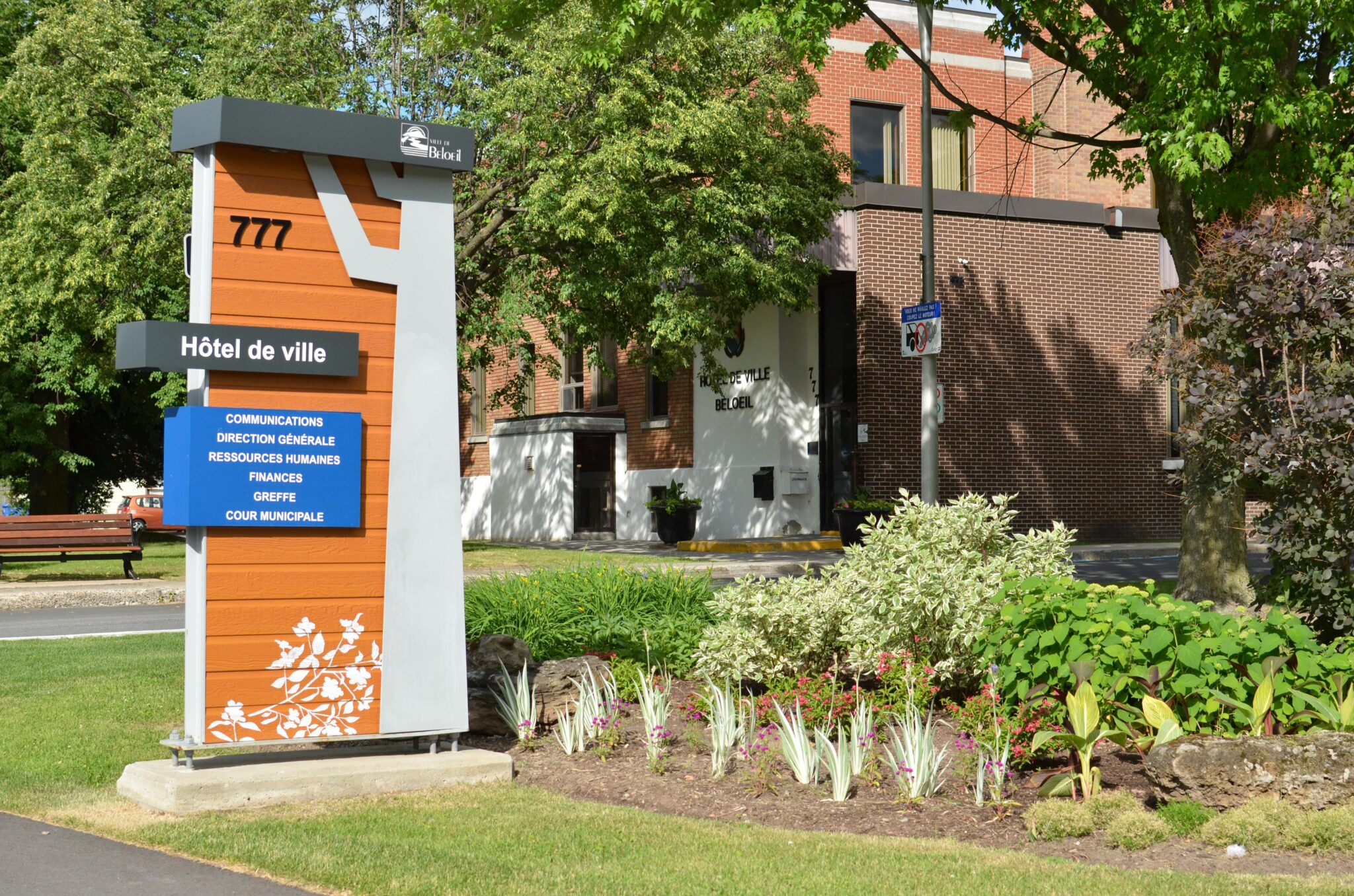 Façade de l’hôtel de ville de Beloeil avec son enseigne moderne orange et grise indiquant les services municipaux, entourée de végétation et d’un aménagement paysager fleuri.