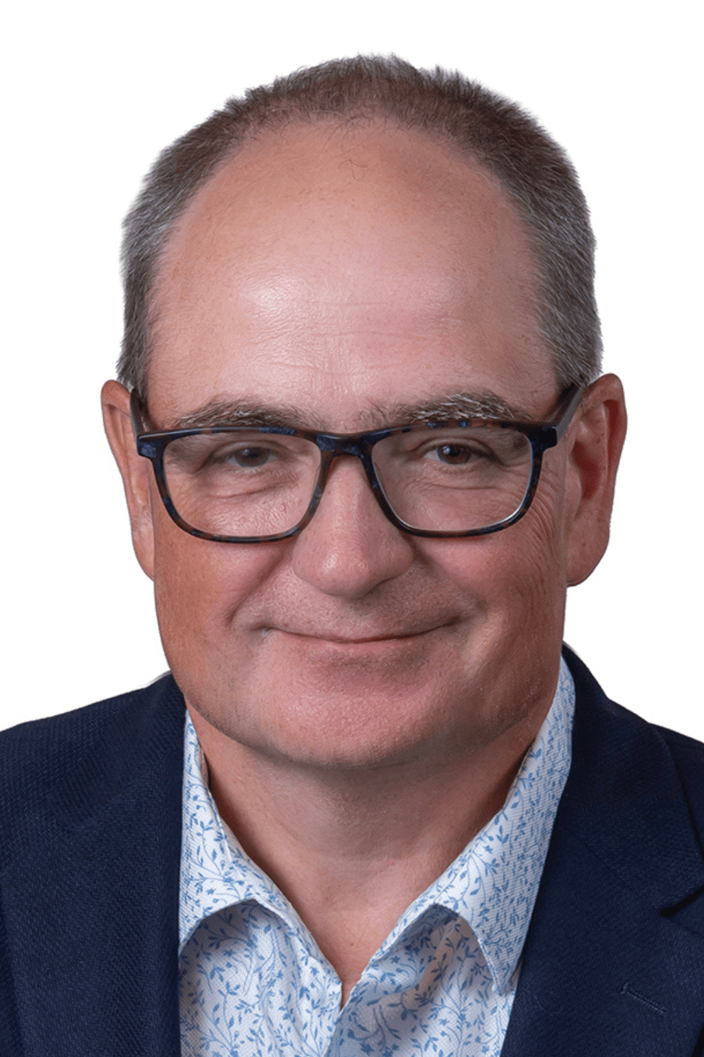 Portrait d’un homme portant des lunettes noires, une chemise pâle à motifs et un veston bleu foncé, souriant légèrement devant un fond blanc.