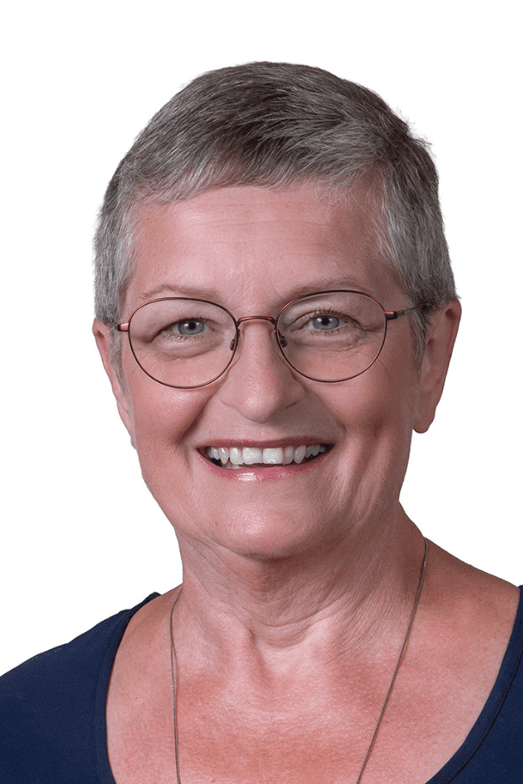 Portrait d’une femme portant des lunettes rondes, des cheveux courts gris et un haut bleu foncé, souriante devant un fond blanc.
