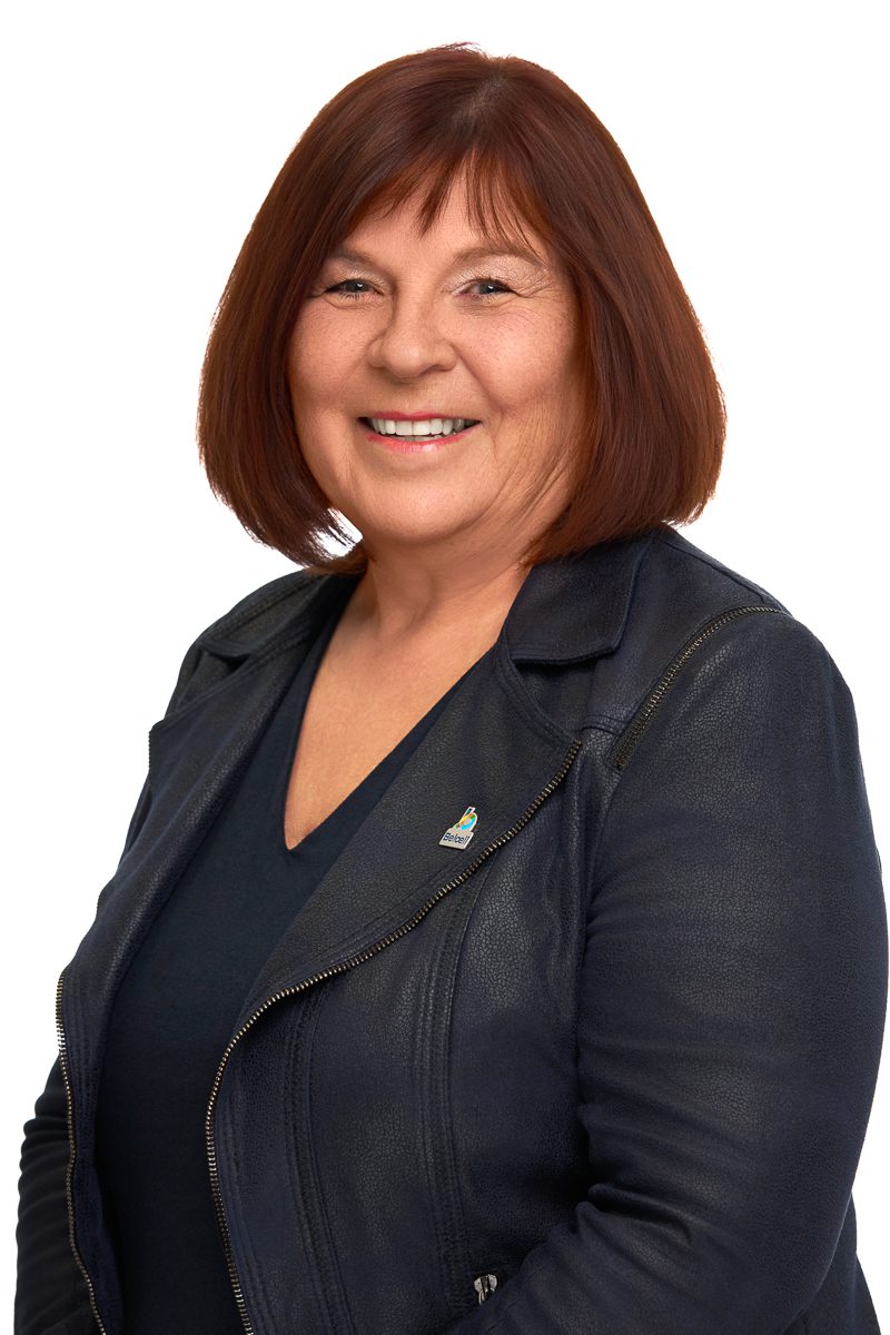 Portrait d’une femme souriante aux cheveux bruns coupés au carré, portant un haut foncé et une veste en cuir bleu foncé, sur fond blanc.