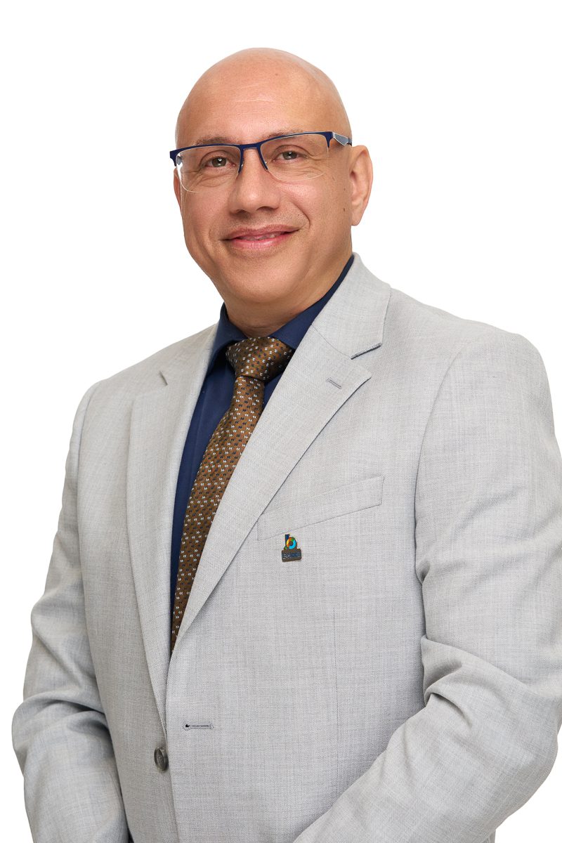 Portrait d’un homme chauve souriant, portant des lunettes à monture bleue, un veston gris pâle, une chemise bleu foncé et une cravate brune à motifs, sur fond blanc.