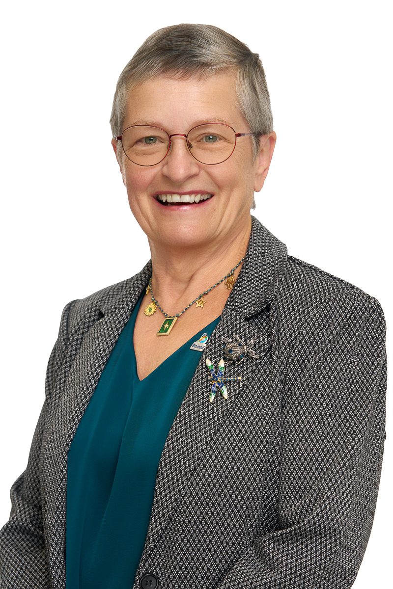 Portrait professionnel d’une femme souriante, aux cheveux gris courts, portant des lunettes rondes, un chemisier turquoise et un veston gris orné de plusieurs broches décoratives.