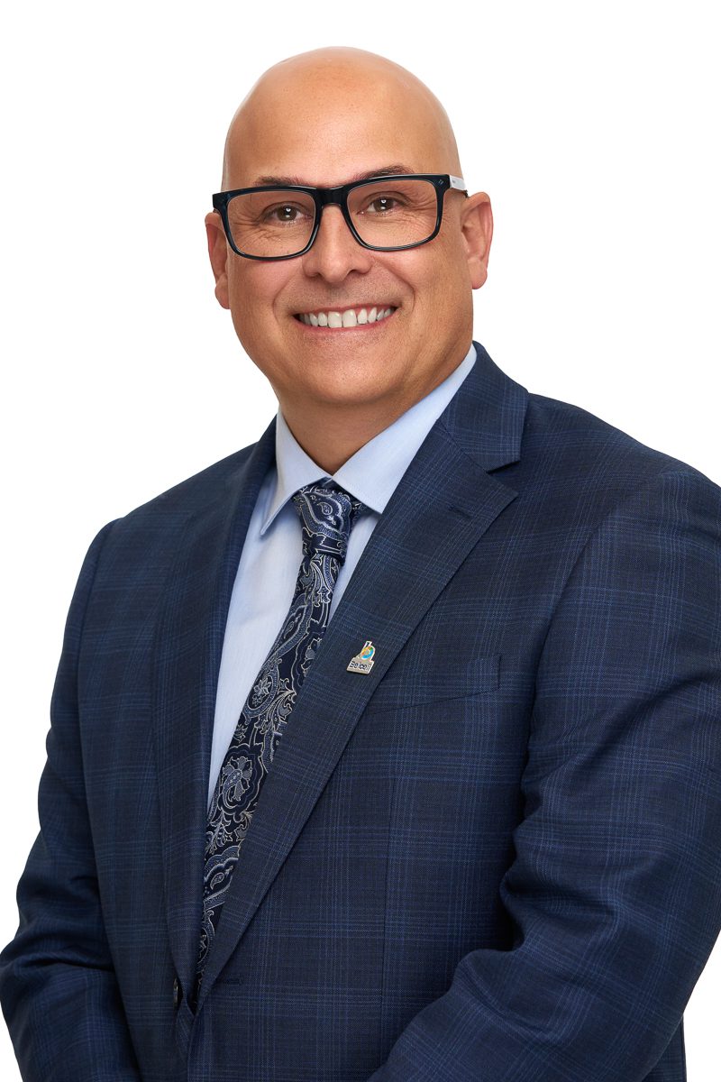 Portrait d’un homme chauve souriant portant des lunettes noires, un veston bleu foncé à motif discret, une chemise bleu pâle et une cravate à motifs paisley, sur fond blanc.