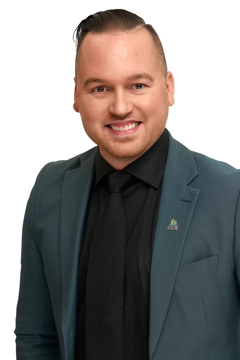Portrait d’un homme souriant portant un veston vert foncé, une chemise noire et une cravate noire texturée, sur fond blanc.