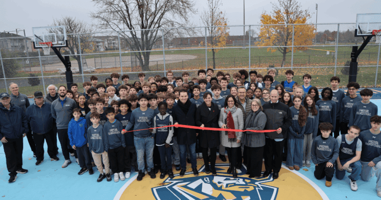 « Grand groupe de personnes rassemblées sur un terrain de basketball extérieur pour une cérémonie d’inauguration. Plusieurs tiennent un ruban rouge au centre du terrain. Deux paniers de basketball sont visibles, et des arbres aux couleurs automnales se trouvent en arrière-plan. »