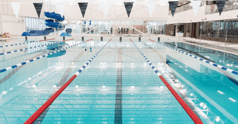 « Piscine intérieure avec plusieurs couloirs de nage séparés par des lignes flottantes. Des fanions bleus et blancs sont suspendus au-dessus de l’eau. À l’arrière-plan, une grande structure de glissades bleues est visible. »