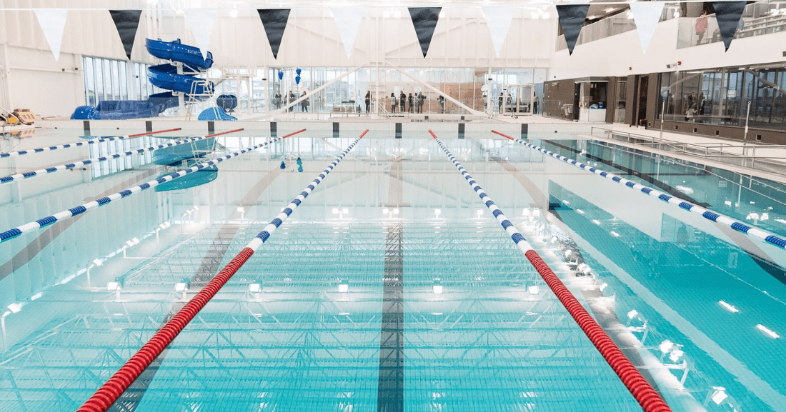 « Piscine intérieure avec plusieurs couloirs de nage séparés par des lignes flottantes. Des fanions bleus et blancs sont suspendus au-dessus de l’eau. À l’arrière-plan, une grande structure de glissades bleues est visible. »