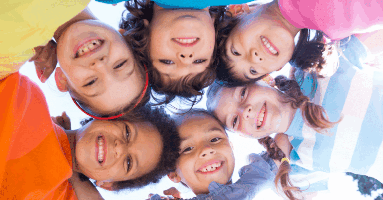 « Six enfants souriants, debout en cercle, regardant vers la caméra placée au sol, sous un ciel bleu dégagé. »