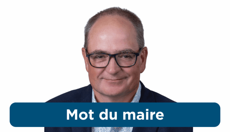 Portrait d’un homme d’âge moyen portant des lunettes, un veston bleu et une chemise pâle à motifs, souriant légèrement devant un fond blanc.