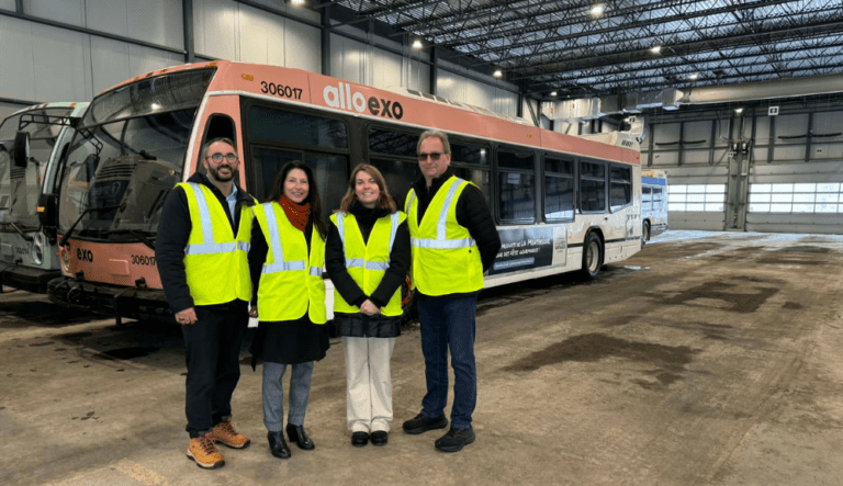 Quatre adultes portant des vestes de sécurité jaunes se tiennent debout dans un garage industriel, devant un autobus de transport collectif Allo exo.
