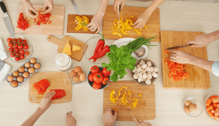 Vue du dessus de plusieurs personnes découpant des légumes colorés (poivrons, tomates, champignons) sur des planches à découper, autour d’une table de cuisine.