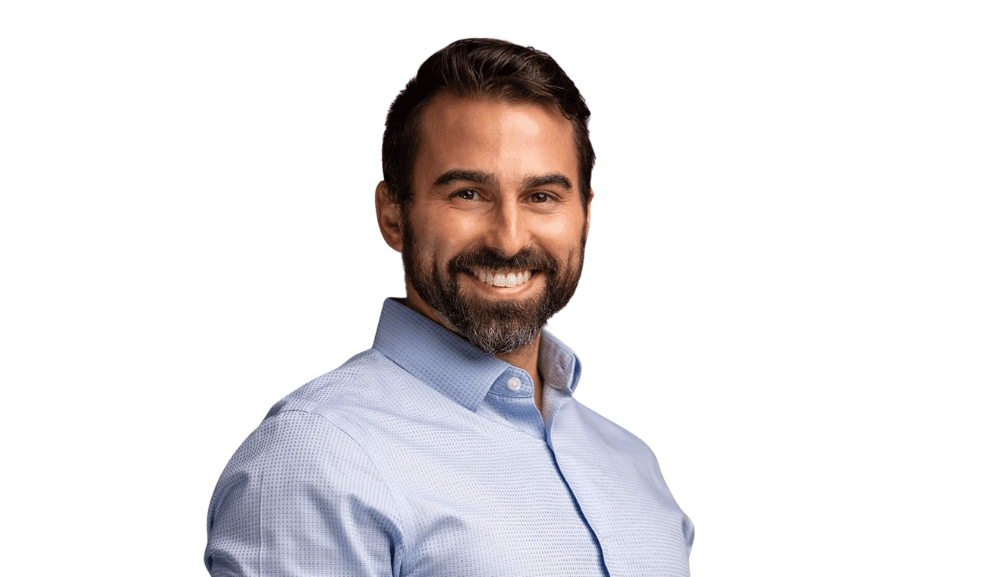 Portrait d’un homme souriant, aux cheveux foncés et à la barbe, portant une chemise bleu pâle sur fond gris clair.
