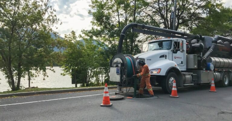 Opération de nettoyage d’égout avec camion aspirateur et cônes de signalisation sur une rue bordée d’arbres près d’un plan d’eau.
