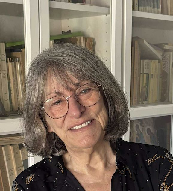 Portrait d’une personne portant des lunettes, assise devant une bibliothèque vitrée remplie de livres.