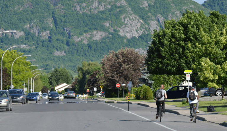 Deux cyclistes roulent sur une bande cyclable en bordure d’une rue résidentielle, avec des montagnes verdoyantes en arrière-plan.
