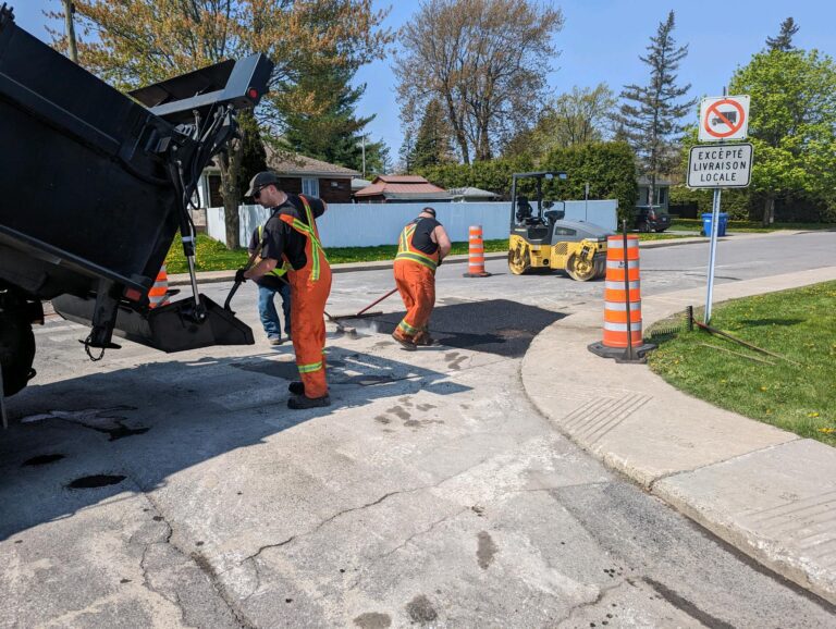 Travaux de réparation de chaussée dans une rue résidentielle, avec camion, ouvriers et rouleau compresseur.