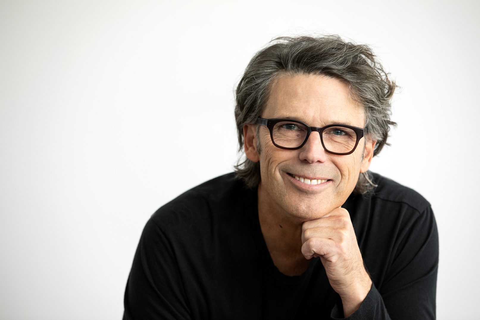 Portrait d’une personne portant des lunettes à monture foncée et un chandail noir, photographiée sur fond gris clair. La personne est légèrement penchée vers l’avant, avec la main appuyée sous le menton, dans une pose professionnelle et épurée.