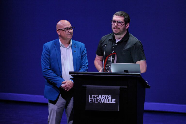 Deux hommes sur une scène, derrière un lutrin portant l’inscription « Les Arts et la Ville »; l’un tient un trophée et prend la parole au micro.
