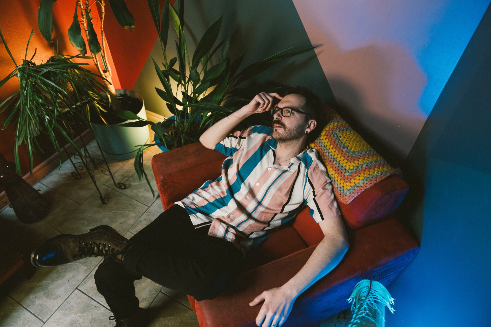 Personne portant des lunettes et une chemise à rayures multicolores, allongée dans un fauteuil rouge à l’intérieur d’une pièce décorée de plusieurs plantes d’intérieur. L’éclairage coloré crée des contrastes orangés, verts et bleus dans l’espace.