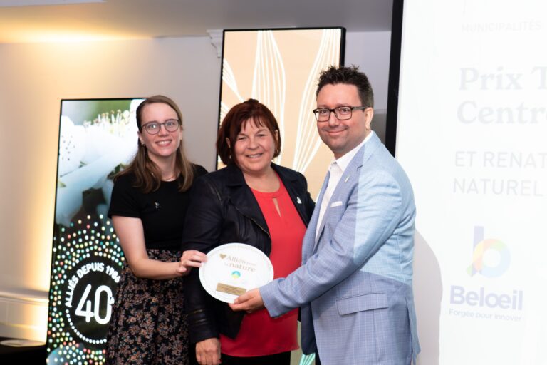 Trois personnes souriantes tiennent une plaque de reconnaissance lors d’une cérémonie soulignant le 40e anniversaire de Nature-Action Québec. Le logo de la Ville de Beloeil apparaît sur l’écran à droite.