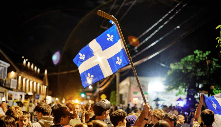 Une foule célèbre dans une rue la nuit tandis qu’une personne brandit un drapeau du Québec fixé à un bâton de hockey.