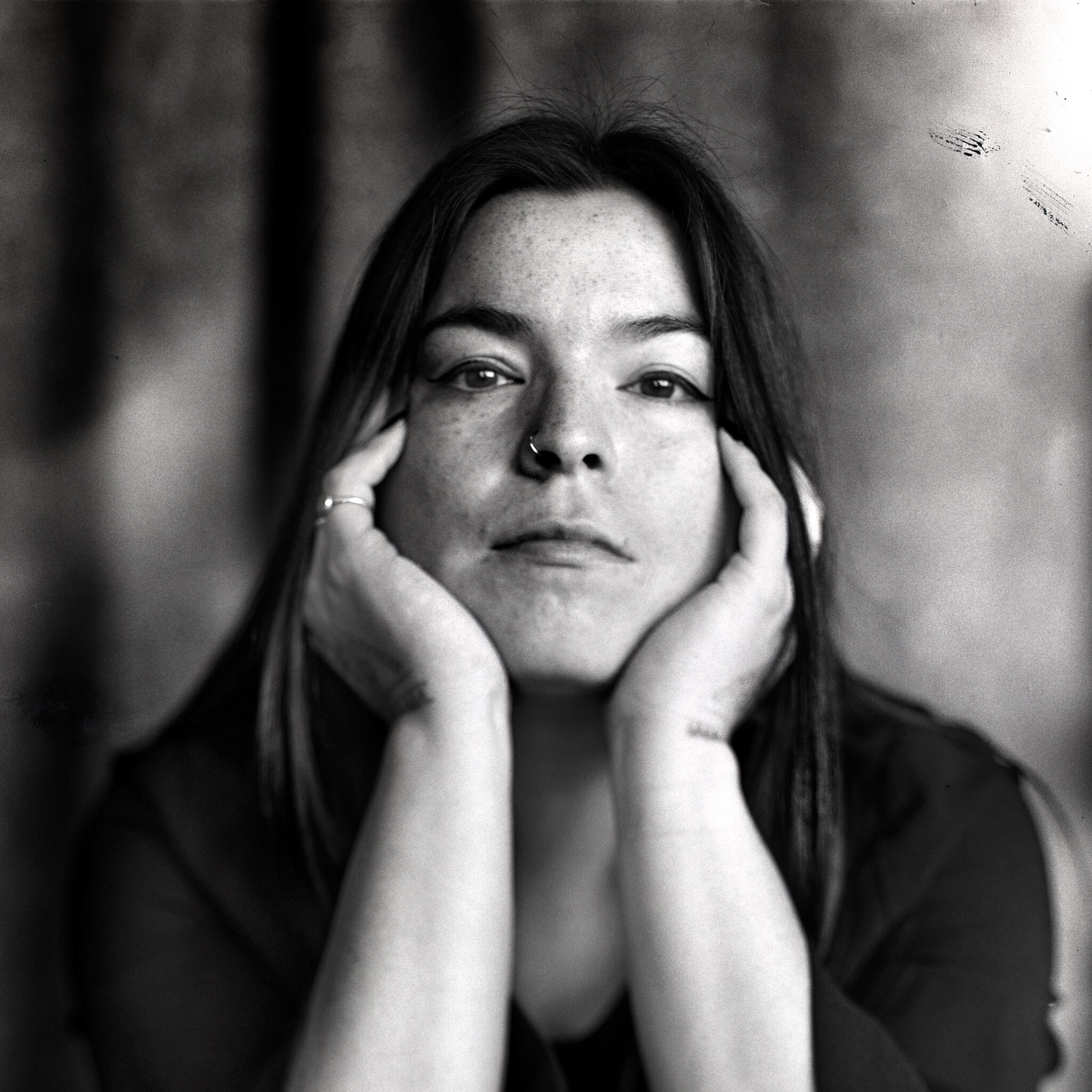 Portrait en noir et blanc d’une personne regardant vers l’objectif, le visage encadré par les deux mains. La personne porte un anneau au nez et une bague à un doigt. L’arrière-plan est flou, ce qui met l’accent sur le visage au centre de l’image.