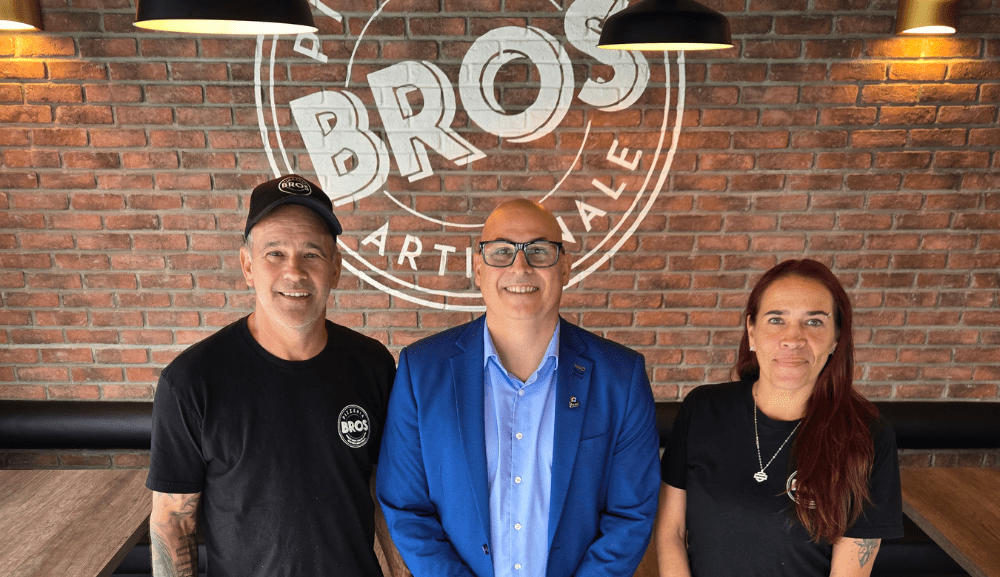 Trois personnes sourient devant un mur de briques affichant le logo « BROS artisanale », dans un décor de restaurant.