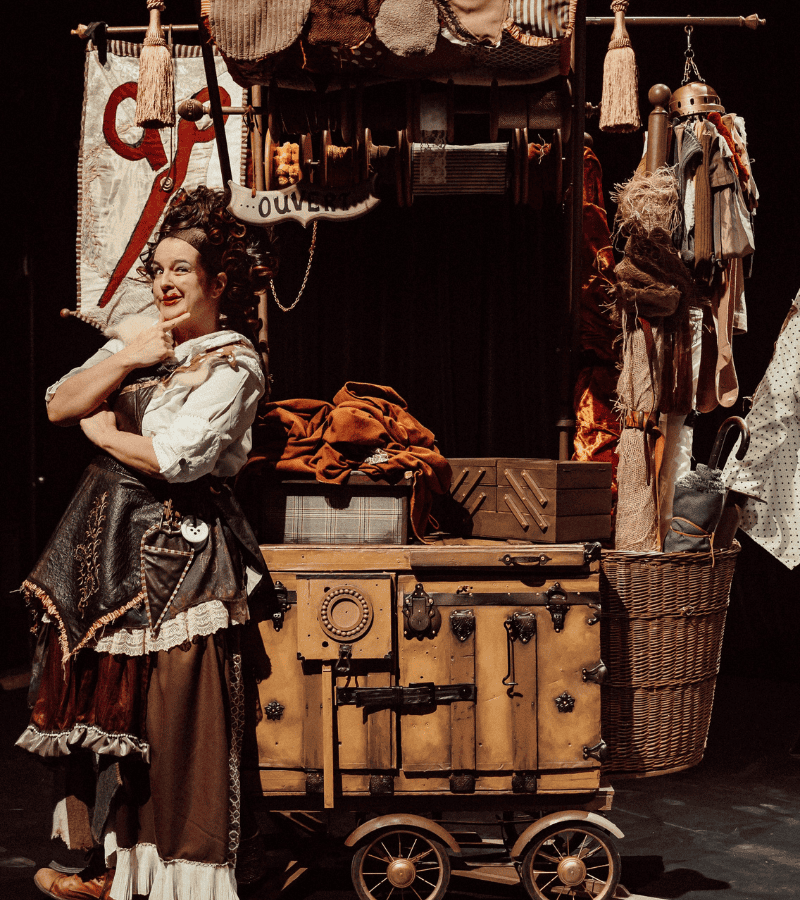 Artiste costumée debout à côté d’un chariot décoré rempli d’objets, de tissus et d’accessoires de spectacle sur une scène.