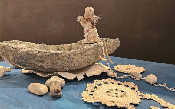 Œuvre artistique représentant une petite embarcation texturée accompagnée d’une figurine, de pierres et d’éléments en crochet rappelant un environnement marin.