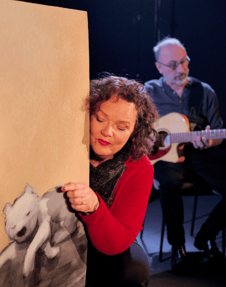 Prestation artistique sur scène combinant musique et création visuelle, avec une personne manipulant un grand panneau illustré et une autre jouant de la guitare à l’arrière-plan.