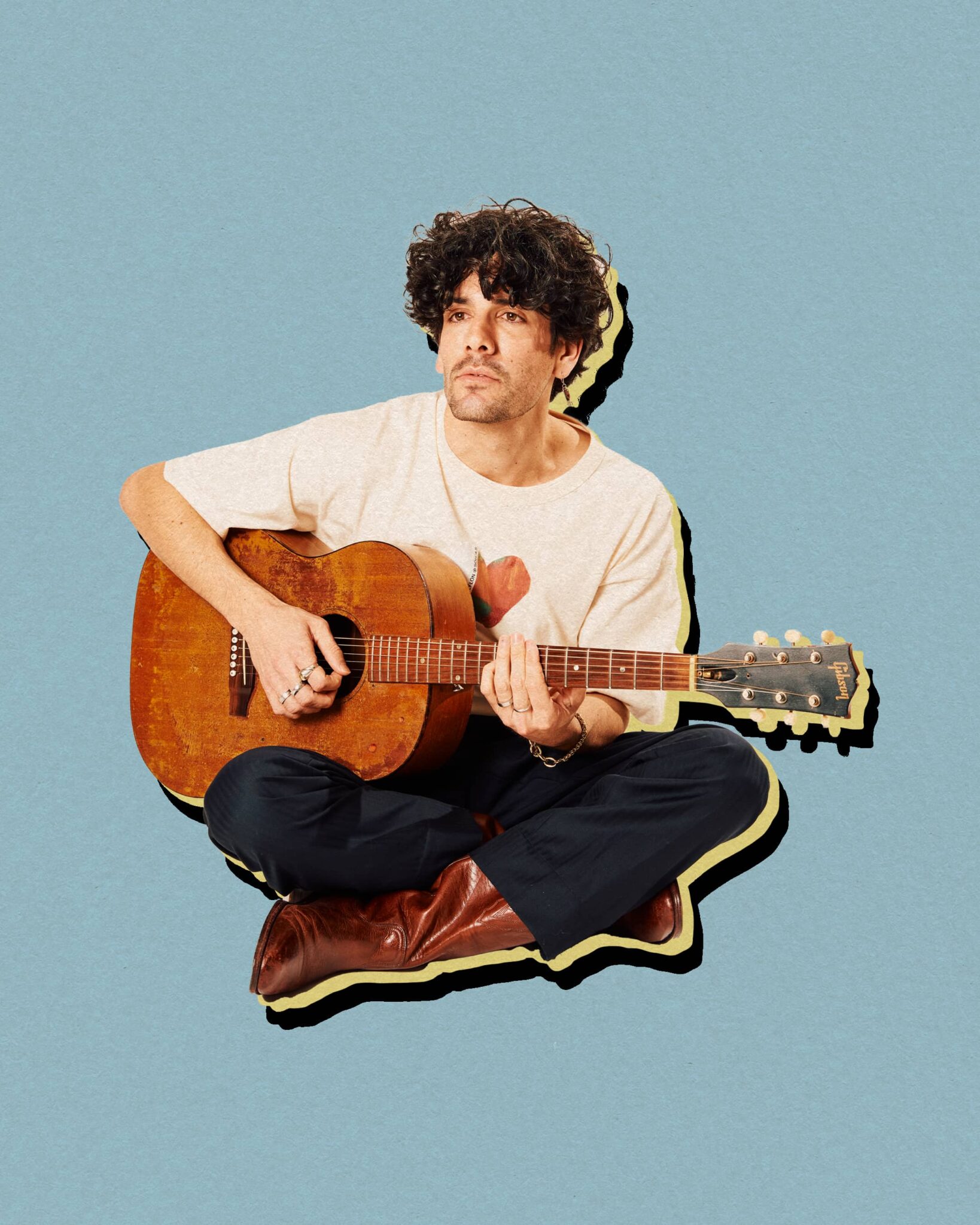 Personne assise au sol avec une guitare acoustique sur les genoux, devant un fond bleu uni.