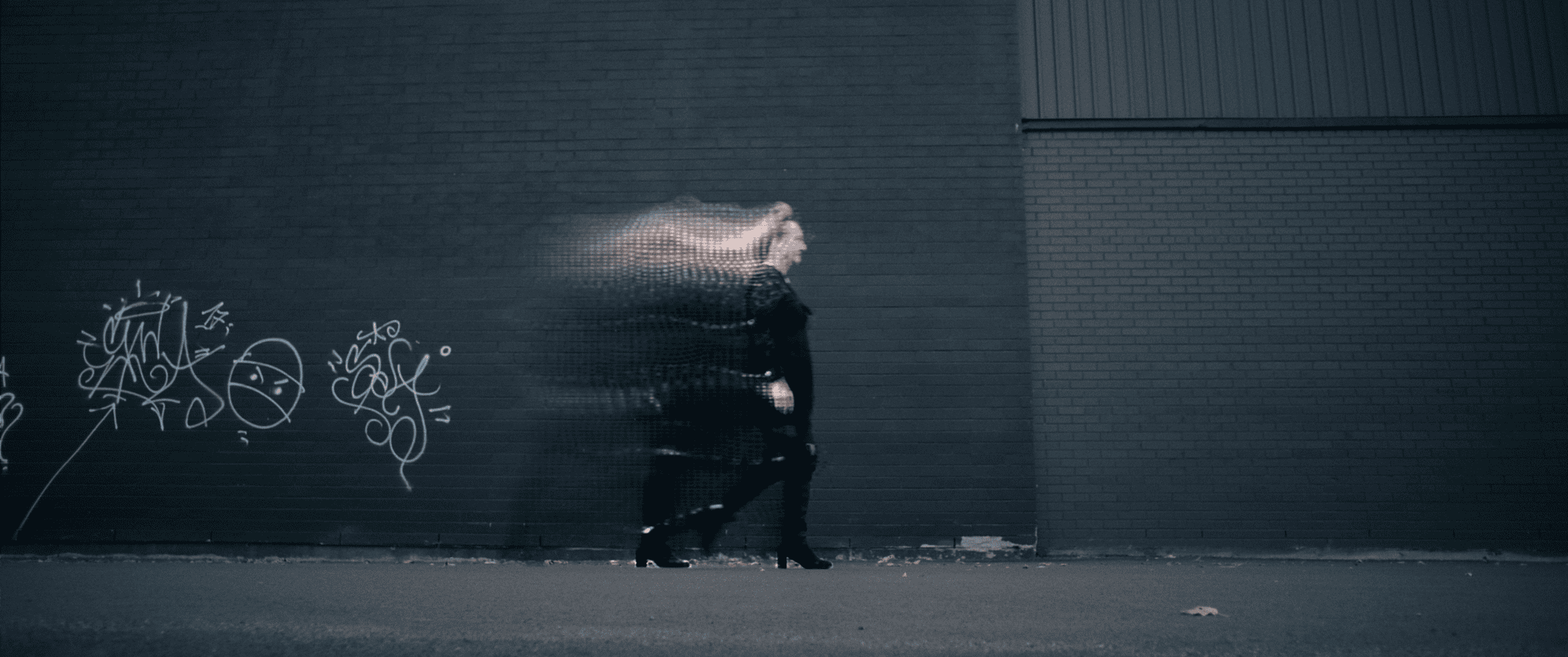 Photographie en mouvement montrant une personne marchant devant un mur de briques sombre couvert de quelques graffitis.