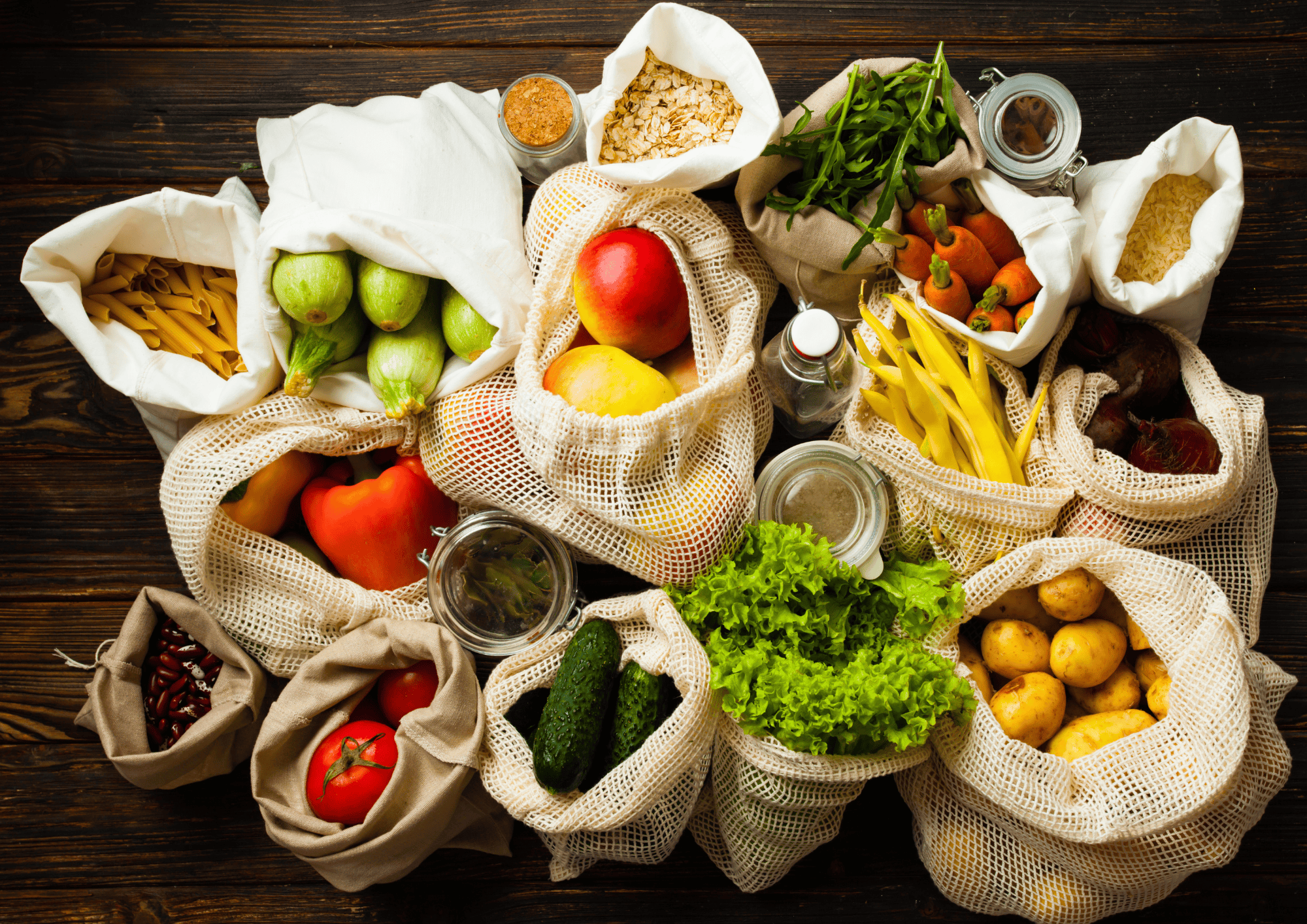 Aliments frais et produits en vrac présentés dans des sacs réutilisables en tissu et en filet sur une table de bois, comprenant des légumes, des fruits, des pâtes, des pommes de terre et des légumineuses.
