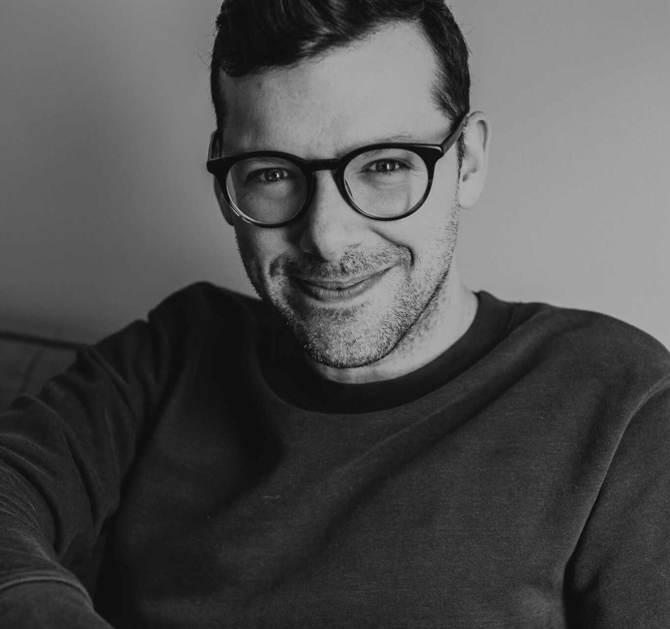Portrait en noir et blanc d’une personne portant des lunettes et un chandail foncé devant un fond uni.