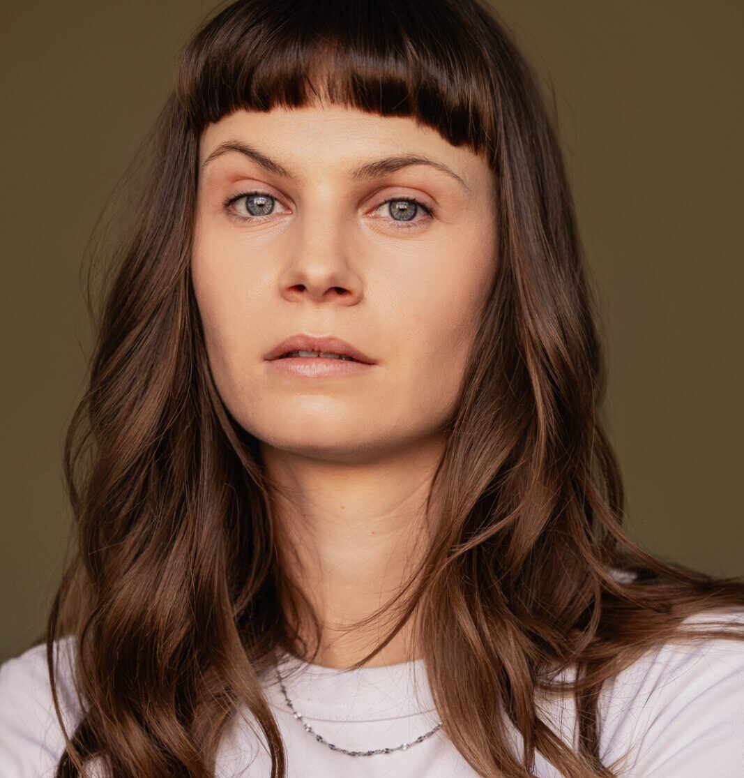 Portrait rapproché d’une personne aux cheveux longs portant un haut clair devant un fond uni.