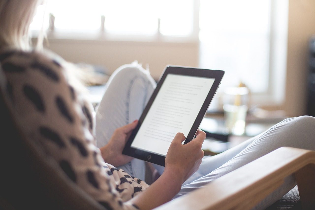 Personne assise dans un fauteuil lisant un document numérique sur une tablette dans une pièce lumineuse.