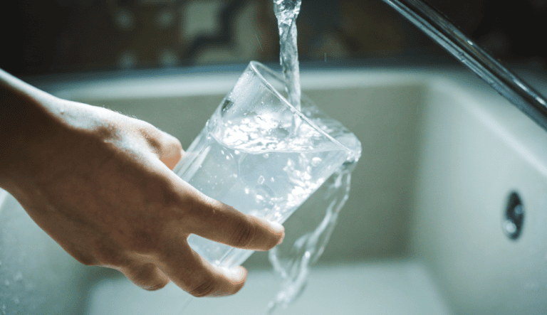 Une main incline un verre transparent sous un robinet pendant que l’eau déborde dans l’évier.
