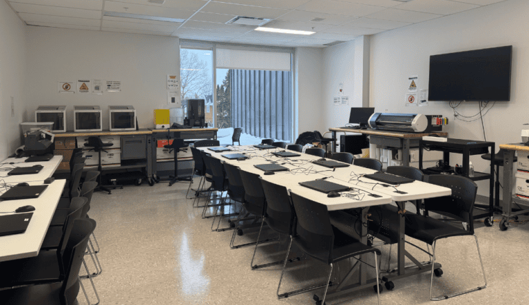 Salle informatique équipée de longues tables, de nombreux ordinateurs portables, de chaises, d’imprimantes et d’un grand écran mural.
