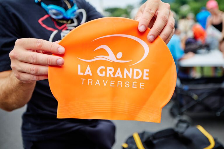 Une personne tient un bonnet de bain orange portant le logo et les mots « La Grande Traversée ».