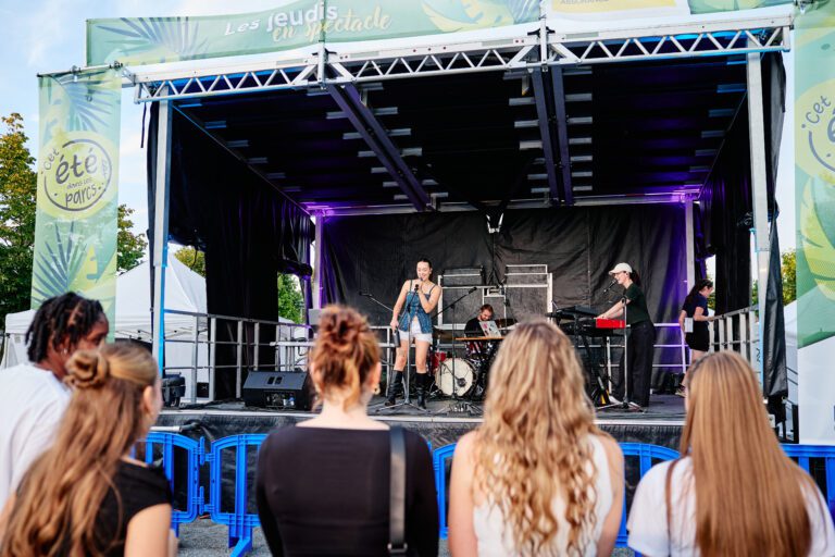 Concert en plein air sur une scène du festival « C’est l’été dans les parcs », devant un public rassemblé derrière des barrières.