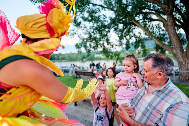 Artiste en costume jaune et rose tendant les mains vers des enfants, tandis qu’un homme tient une fillette dans ses bras lors d’une activité extérieure.