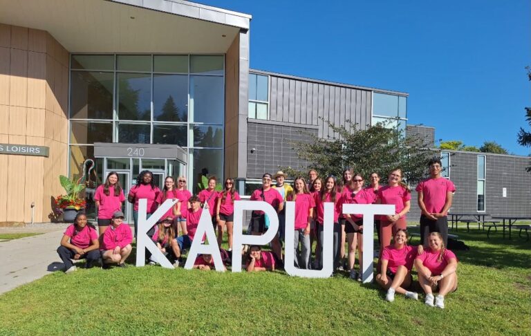 Groupe de personnes en chandails roses posant devant de grandes lettres blanches formant « KAPUT », à l’extérieur d’un centre de loisirs.