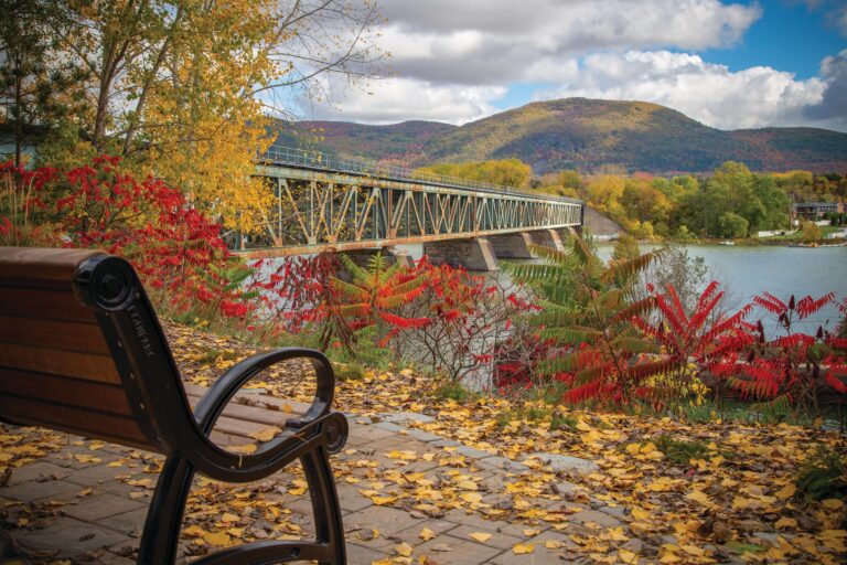 Banc au bord de l’eau face à un long pont métallique, avec des montagnes couvertes de couleurs automnales en arrière-plan.