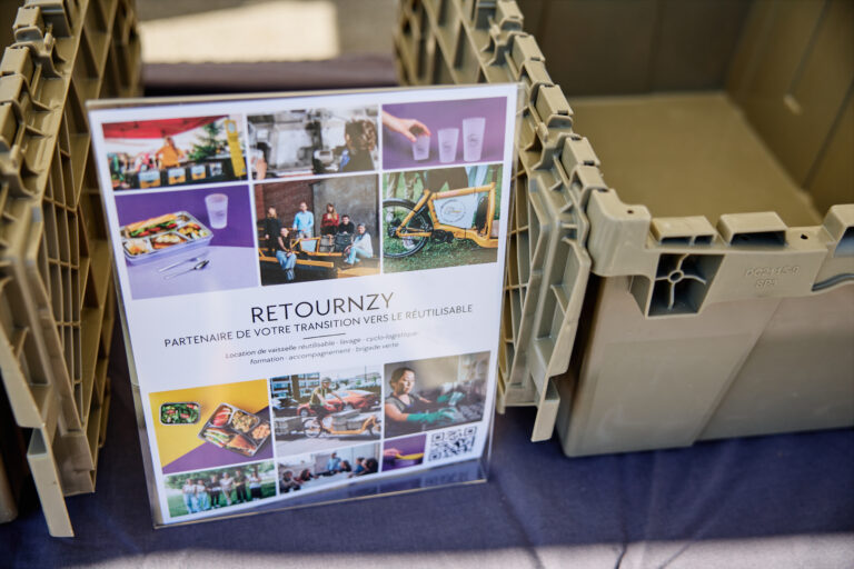 Bacs de transport réutilisables gris disposés autour d’une affiche de l’entreprise Retournzy, qui présente ses services liés à la vaisselle réutilisable et à la cyclologistique.