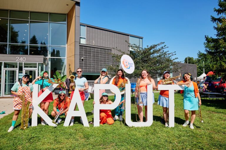 Groupe de musiciennes posant avec des saxophones, trombones et autres cuivres derrière de grandes lettres blanches formant « KAPUT », devant un bâtiment moderne.